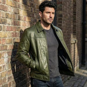 May include: An olive green leather jacket with a classic collar, a full-length front zipper, and two side pockets. The jacket is worn over a plain dark gray t-shirt and paired with dark blue denim jeans, showcasing a smooth, polished finish.