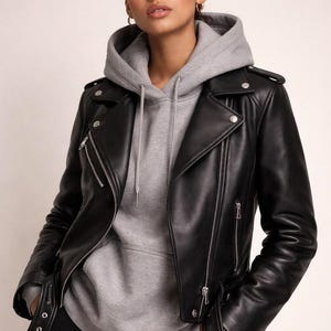 Peut inclure: Une veste de moto en cuir noir avec des fermetures éclair, des boutons-pression et des épaulettes en métal argenté, portée sur un sweat à capuche gris avec un cordon de serrage. La veste présente un large revers cranté et une ceinture.