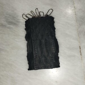 Puede incluir: Un estuche para llaves rectangular de color negro con un patrón texturizado similar a escamas en su superficie exterior. El borde superior del estuche cuenta con seis clips metálicos plateados para sujetar llaves, alineados a lo largo.