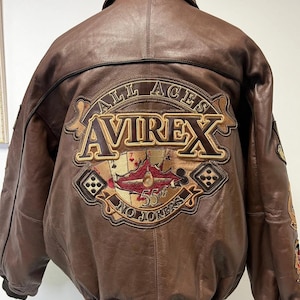 Könnte beinhalten: Braune Leder-Bomberjacke mit großem gesticktem Aufnäher auf dem Rücken. Der Aufnäher zeigt den Text "ALL ACES AVIREX 55th NO JOKERS" mit Spielkarten- und Würfelmotiven. Die Jacke hat einen gerippten Kragen und Bündchen.