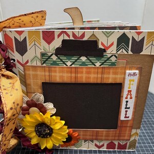 Peut inclure: Un mini-album artisanal sur le thème de l'automne avec un accent en bois en forme de citrouille. L'album présente du papier à motifs aux couleurs automnales, un cadre à carreaux et une décoration de tournesol. Le texte "Hello FALL" est visible.