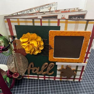 Könnte beinhalten: Ein Scrapbook mit Herbstthema. Der Umschlag zeigt ein Karomuster, eine gelbe Blume, ein braunes Blatt und das Wort "Herbst" in braunen Buchstaben. Es gibt einen kleinen braunen Rahmen mit orangefarbenem Rand.