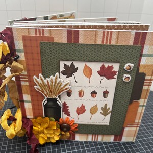 Pode incluir: Um álbum de scrapbook feito à mão com tema de outono. O álbum apresenta papel estampado em laranja, marrom e verde. Os enfeites incluem adesivos de folhas e bolotas, um vaso de trigo e fitas e flores decorativas. O álbum é encadernado com um anel de metal preto.