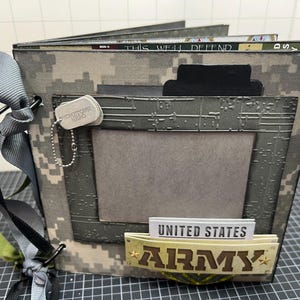 May include: A handmade scrapbook with a camouflage pattern cover. The cover features a metal dog tag, a decorative frame, and a banner that reads "United States Army". The pages are visible and the book is tied with a gray and green ribbon.