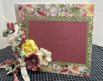 May include: A pink scrapbook with a floral design and a green lace border. The scrapbook has a pink paper insert and is decorated with yellow, white, and pink flowers.