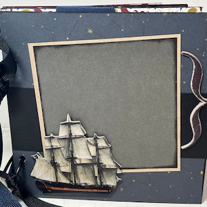 Op de afbeelding: Een marineblauw scrapbook met een zwart-witte illustratie van een schip op de voorkant. De omslag heeft een gouden lijst om een grijs vierkant met een bruine snor aan de rechterkant. Het scrapbook is vastgebonden met een zwart lint.