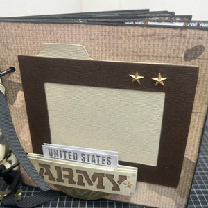 May include: A brown scrapbook with a camouflage pattern and a large rectangular window. The scrapbook has a black ribbon and a gold star on the front. The text "UNITED STATES ARMY" is visible on the front of the scrapbook.