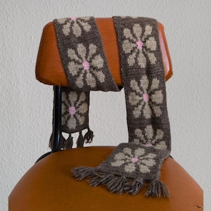 May include: A brown crocheted scarf with a floral pattern in tan and pink. The scarf is draped over the back of an orange chair. The scarf has tassels at the ends.