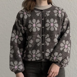 May include: A button-up cardigan sweater in a dark gray color with a floral pattern in light gray and pink. The sweater has long sleeves and a round neckline. The floral design is repeated across the entire sweater.