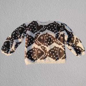 Pode incluir: Um suéter creme com um padrão repetido de peixes marrons escuros. O tricô de manga comprida tem gola redonda e punhos e bainha canelados. O desenho do peixe é detalhado com pequenos pontos brancos.