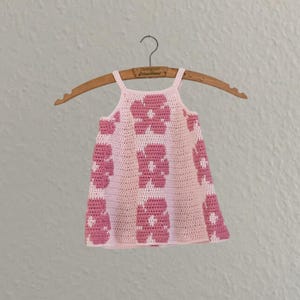 Peut inclure: Une robe en crochet rose clair avec un motif floral rose foncé. La robe est présentée sur un cintre en bois. La robe a un design sans manches avec de fines bretelles.