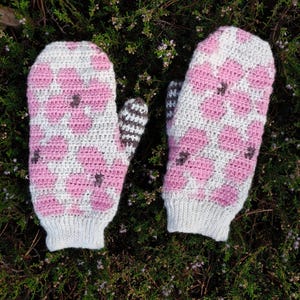 May include: A pair of handmade mittens with a floral pattern. The mittens feature a pink and white floral design on the front and back, with a gray cuff and a brown and white striped thumb. The mittens are lying on a bed of green foliage.