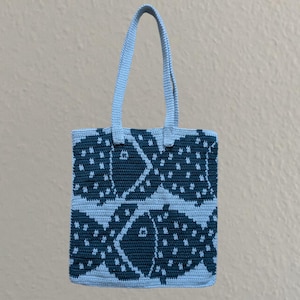 May include: A light blue crochet tote bag featuring a repeating pattern of dark blue fish. The bag has a rectangular shape with a matching light blue handle. The fish pattern is detailed with white dots.