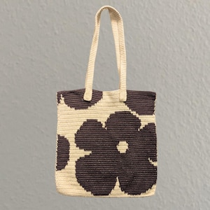 Puede incluir: Un bolso tote de crochet color crema con un patrón repetido de grandes formas de flores marrón oscuro. El bolso tiene dos asas color crema y está diseñado para transportar objetos. El bolso está hecho de un material tejido.