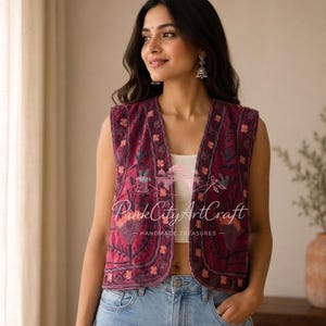 May include: A sleeveless burgundy waistcoat featuring intricate floral embroidery in shades of pink, orange, and teal. The garment has an open front design with a rounded neckline and is layered over a white top and light blue denim jeans.