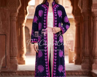 May include: A long, dark navy blue velvet coat featuring vibrant purple floral embroidery and a bright pink trim. The coat has a high collar and a repeating heart-shaped pattern along the front opening. It is worn over a white top and white trousers.