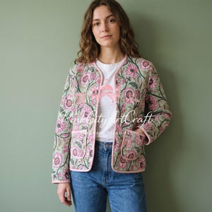 May include: A quilted jacket featuring a light green base with a pink and dark brown floral pattern, long sleeves, two front pockets, and pink trim. The jacket is worn over a white t-shirt and blue jeans. The text PinkCityArtCraft is visible.