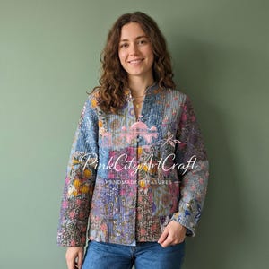 Op de afbeelding: Een gewatteerde jas met een bloemen patchwork design in blauw, roze en geel. De jas heeft een opstaande kraag en lange mouwen. De tekst "Pink City Art Craft - Handmade Treasures" is zichtbaar op de voorkant.