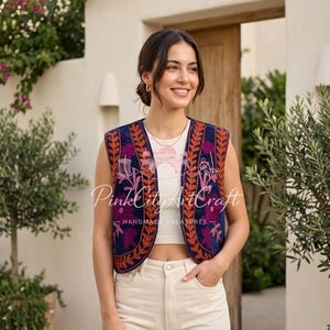 May include: A sleeveless navy blue waistcoat featuring vibrant orange and purple floral embroidery along the front edges. The waistcoat is worn over a white ribbed tank top and cream-coloured trousers. The text PinkCityArtCraft is visible on the waistcoat.