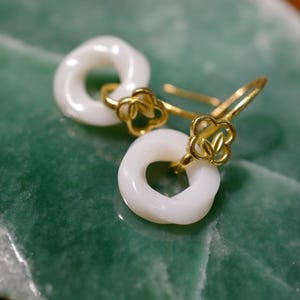 Puede incluir: Un par de pendientes blancos y circulares con detalles dorados. Los pendientes se exhiben sobre una superficie verde y texturizada. Los pendientes tienen un pequeño diseño floral donde los ganchos se conectan a la parte circular.