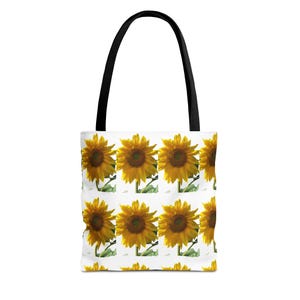 May include: A white tote bag featuring a repeating pattern of vibrant yellow sunflowers with dark brown centers and green stems. The bag has two sturdy black shoulder straps attached to the top, providing a clear contrast against the white fabric.