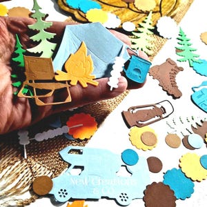 May include: A collection of camping-themed paper die-cuts including a light blue tent, a yellow campfire, a brown folding chair, a blue mug, a brown lantern, a brown hiking boot, green pine trees, and a light blue camper with the text New Creations & Co.