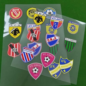 May include: A collection of colorful soccer team logo stickers, featuring various shield and circular designs. The stickers include team names like "FC Lahti," "Atlante," and "A.E.K." Some logos incorporate soccer balls and the year "1996."