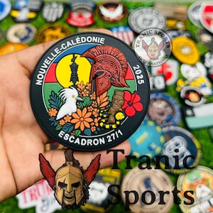 May include: A round black patch with the text "NOUVELLE-CALÉDONIE" and "ESCADRON 27/1". The patch features a red helmet, a white bird, a yellow sun, and a tribal figure. The year "2025" is also displayed. The patch is held in a hand.