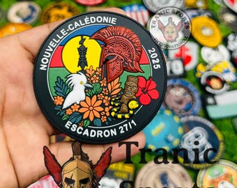 May include: A round black patch with the text "NOUVELLE-CALÉDONIE" and "ESCADRON 27/1". The patch features a red helmet, a white bird, a yellow sun, and a tribal figure. The year "2025" is also displayed. The patch is held in a hand.