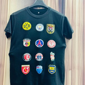 Peut inclure: T-shirt noir à col rond orné de nombreux écussons brodés. Les écussons affichent des logos d'équipes et des textes tels que "BVB 09", "Manchester City", "Guardia Civil", "Fortuna Koln", "Charlton Athletic", "Washington Wizards", "Degerfors", "CK Potop", et "NSK 1952".
