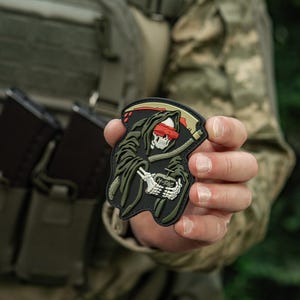 May include: A black, green, and red patch featuring a Grim Reaper design. The Reaper is depicted with a skull, blindfold, and scythe. The patch includes the text "M-TAC" and is held in a person's hand.