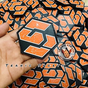 Könnte beinhalten: Haufenweise sechseckige schwarze und orangefarbene Aufnäher mit der Zahl "51" in einem stilisierten Design. Die Aufnäher sind zusammengelegt, einer wird in der Hand gehalten. Der Text "TRANIC SPORTS" ist sichtbar.