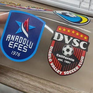 May include: Two shield-shaped stickers. One is blue with white text reading "ANADOLU EFES 1976" and a star logo. The other is red and black with "DVSC" and "Diablo Valley Soccer Club" text, plus a soccer ball graphic.