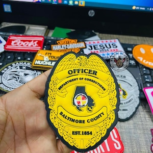 Può includere: Un distintivo da ufficiale della contea di Baltimora giallo e nero con la scritta "OFFICER" e "DEPARTMENT OF CORRECTIONS". Il distintivo presenta il contorno dello stato del Maryland e la scritta "EST.1854". Altre toppe sono visibili sullo sfondo.