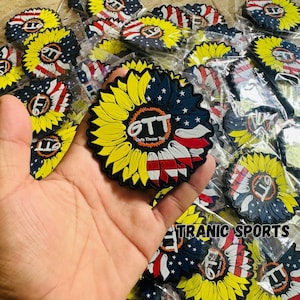 Puede incluir: Una colección de parches con forma de girasol. Cada parche presenta un diseño de girasol amarillo, una sección azul con estrellas blancas y una sección a rayas rojas y blancas, similar a la bandera estadounidense. El texto "GTT Girls Throw Too" es visible.
