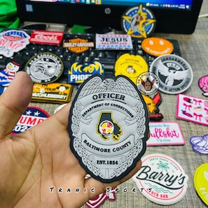 May include: A selection of embroidered patches with diverse designs, including a police badge, a smiley face, and text that reads "JESUS". The patches are in various shapes and colours, with text and graphics.