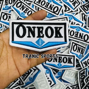 Può includere: Toppa ricamata con la scritta "ONEOK" in lettere nere su sfondo bianco, incorniciata da un bordo nero. Un diamante blu e dettagli blu sono presenti. Il testo "TRANIC SPORTS" è in basso.