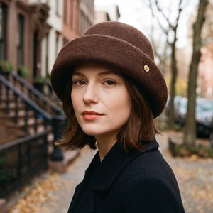 May include: A dark brown felt cloche hat with a rounded crown and a wide, turned-up brim. The hat features a small, circular gold-toned metal accent on the side of the brim. The material has a soft, textured finish consistent with felted wool.