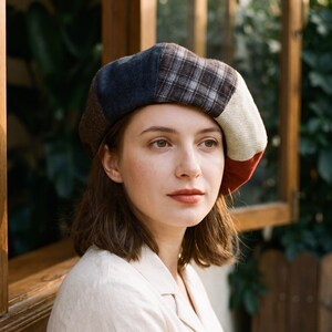 Op de afbeelding: Een veelkleurige patchwork baret met panelen van donkerblauwe, bruine, zwart-wit geruite, crèmekleurige en roestrode stof. De muts is gemaakt van diverse gestructureerde materialen, waaronder wolachtige weefsels en effen gekleurde stofsegmenten.