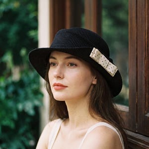May include: A black crocheted hat with a structured brim and a cream-colored ribbon accent featuring small black polka dots. The hat has a textured, woven appearance and is adorned with a decorative bow detail on the side of the crown.