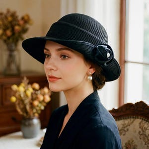 May include: A black wide-brimmed hat with a structured crown and a horizontal ribbed texture. The hat features a decorative black fabric rose accent on the side, which is centered with a single white pearl-like bead for a classic, refined appearance.