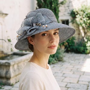 May include: A dark gray straw hat with a wide brim, featuring a large, layered organza flower decoration on the side. The floral accent is crafted from sheer, grid-patterned fabric and is embellished with small, round, cream-colored faux pearls.