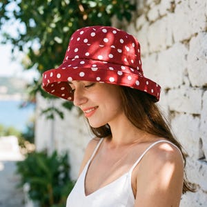 Peut inclure: Un chapeau bob rouge orné d'un motif répétitif à pois blancs. Le chapeau présente un bord large incliné vers le bas et une calotte structurée. Le tissu affiche un imprimé circulaire uniforme sur toute la surface extérieure du couvre-chef.