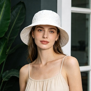 May include: A white, wide-brimmed sun hat with an embroidered floral pattern. The hat has a rounded crown and a broad, gently sloping brim. The hat is made of a lightweight fabric, perfect for sun protection. The hat is worn by a person wearing a beige spaghetti strap top.