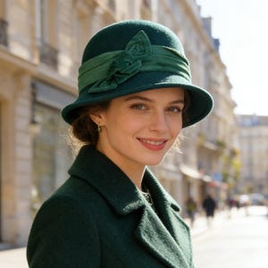 May include: A woman wearing a dark green cloche hat with a matching ribbon and bow detail. She is also wearing a dark green coat. The hat and coat create a vintage, classic look. The background shows a European street scene.
