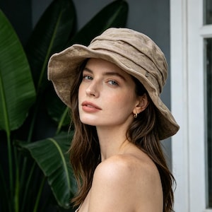 May include: A beige sun hat with a wide brim and a gathered crown. The hat is made of a soft, textured fabric. The hat is worn by a person with long brown hair and gold hoop earrings. The background includes large green leaves and a window.