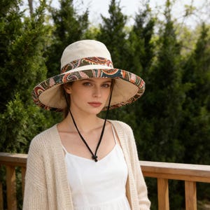 Puede incluir: Un sombrero para el sol color crema con ala ancha y una banda con estampado de cachemira. El sombrero tiene una correa de barbilla negra y se usa al aire libre. El fondo presenta árboles verdes y una barandilla de madera.