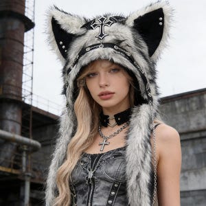 May include: A faux fur hood featuring large animal ears, a black cross emblem, and silver-toned metal chain accents. The hood is patterned in shades of grey, black, and white, with silver studs along the inner ear edges and a black trim border.