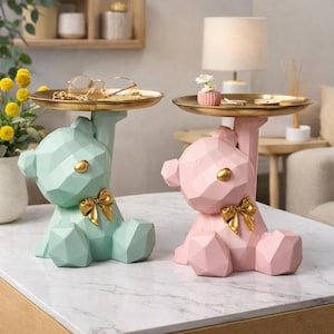 Op de afbeelding: Twee decoratieve berenvormige trays, één mintgroen en één roze, staan op een wit marmeren oppervlak. Elke beer houdt een goudkleurige tray vast. De beren hebben gouden strikken en gouden accenten. Eén tray bevat glazen, de andere een kleine vaas.