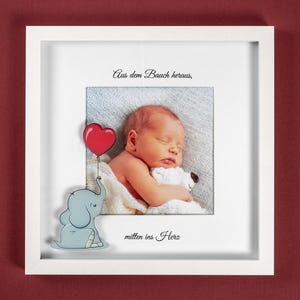Könnte beinhalten: Ein weißer Bilderrahmen mit einem schlafenden Baby, einem roten Herzballon und einem Cartoon-Elefanten. Der Rahmen enthält deutschen Text. Der Hintergrund ist tiefrot.